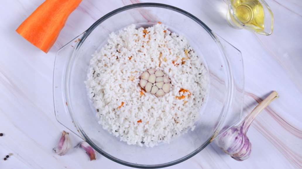 Выложите в форму для запекания зажарку с курицей, сверху добавьте рис и разровняйте по всей поверхности. Вдавите в рис головку чеснока. 