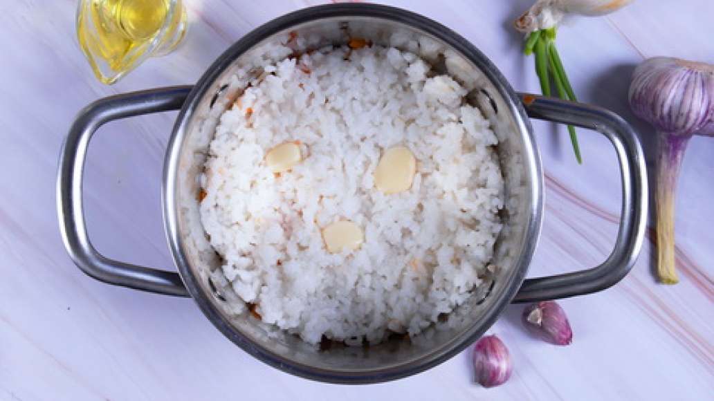 Слейте воду с риса и выложите поверх мясной зажарки. Вдавите в рис очищенные зубчики чеснока. Влейте в кастрюлю горячую воду, накройте крышкой и доведите до кипения. Убавьте огонь и готовьте 25–30 минут, пока вся жидкость не выпарится. 